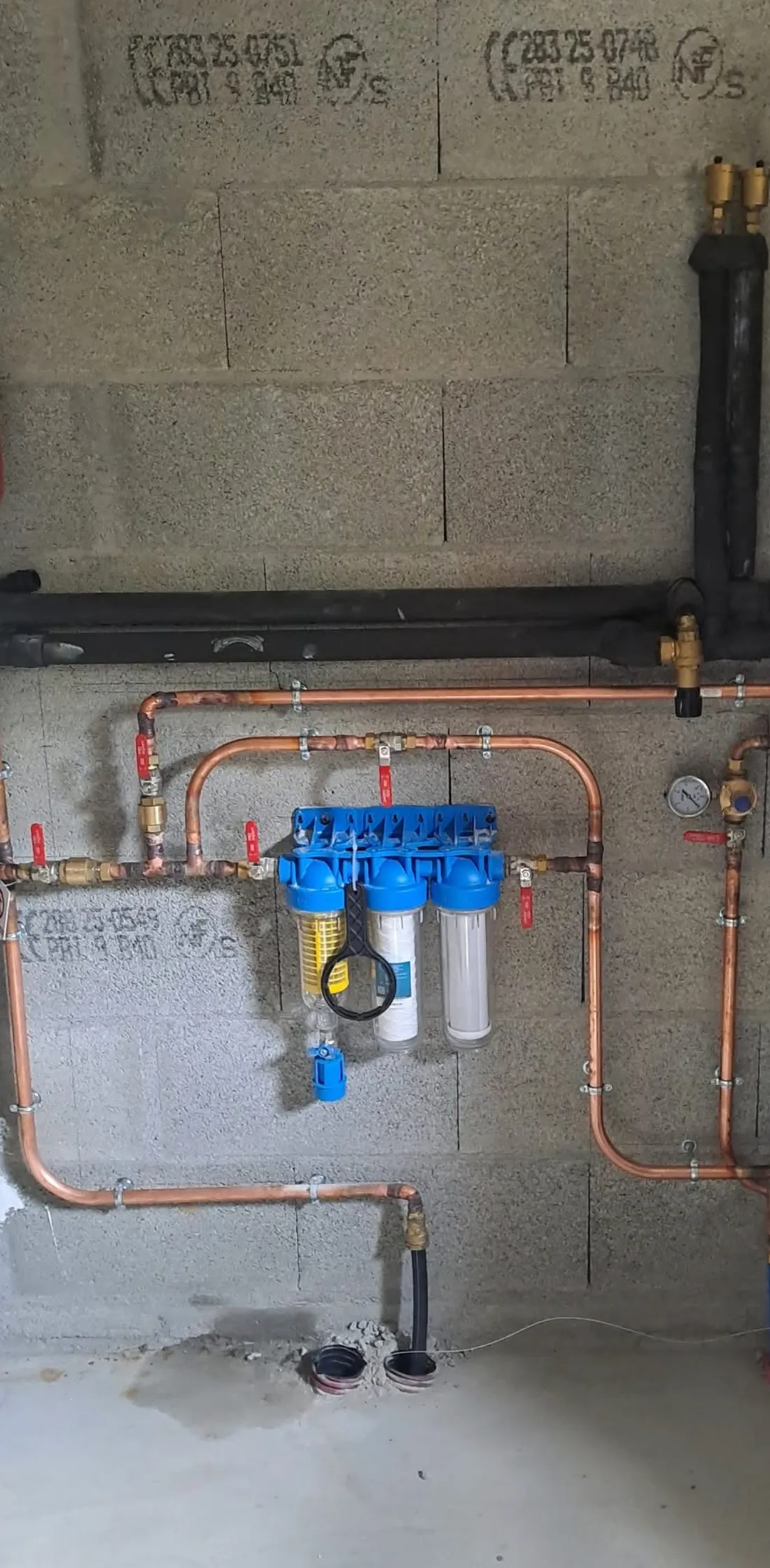 Station de filtration d'eau posée par A2C Plomberie : triple porte-filtre, réducteur de pression et distribution cuivre sur mur de garage